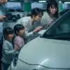 地下駐車場でバッテリーが上がってしまい、エンジンがかからず困惑している日本の家族。スマートキーも反応せず、スマートフォンで対処法を調べている。メーターパネルにはバッテリー警告灯が点灯している。