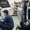 自動車整備工場で、二人の日本人の整備士が新型シエンタと旧型シエンタのオプションカプラー配線図を確認しながら作業している。一人はタブレットで配線図を見ながら、もう一人は懐中電灯で車両内部を照らしている。