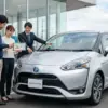 自動車販売店の前で、シエンタNSP170の購入を検討している日本人夫婦と営業担当者が、タブレットを囲んで燃費やリコールの情報を真剣に話し合っている様子。