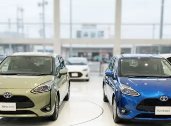 自動車販売店のショールームに並ぶ新型と旧型シエンタ。中央のディスプレイには「3年落ち買取価格 新旧比較 価格変動！」というグラフが表示されており、男女2人の日本人スタッフがそれを見ながら話している。