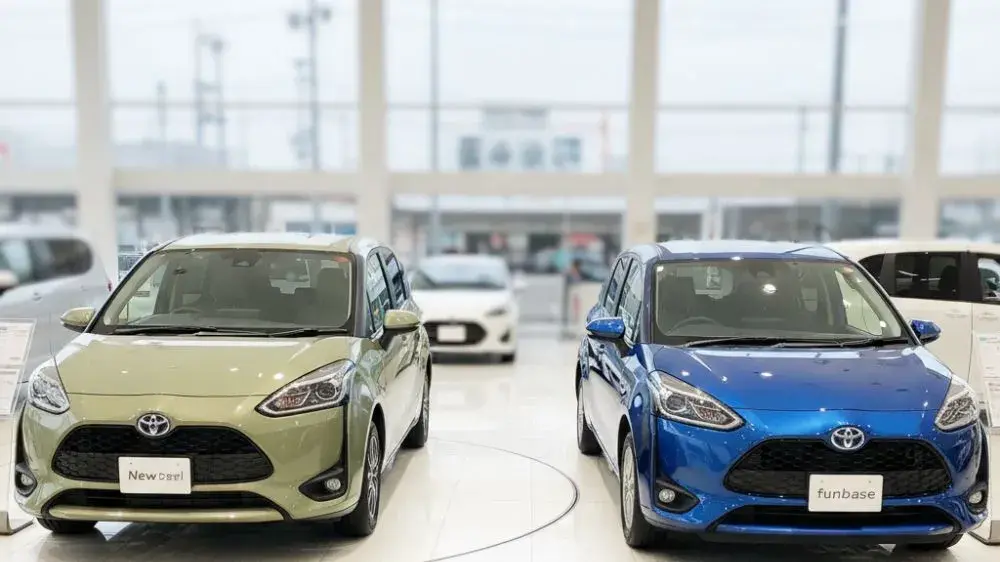 自動車販売店のショールームに並ぶ新型と旧型シエンタ。中央のディスプレイには「3年落ち買取価格 新旧比較 価格変動！」というグラフが表示されており、男女2人の日本人スタッフがそれを見ながら話している。