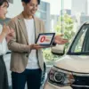 自動車ディーラーでタブレットを使ってカーリースの説明を受け、笑顔で頷く若いカップル。手元には「頭金0円」と表示された画面が見える。