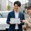 営業担当者がタブレットでカーリースの料金プランを日本人夫婦に説明している様子。白いカーリース車が背景にある。